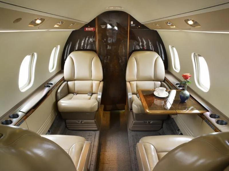 View of a luxurious aircraft cabin interior with tan leather seats, a polished wood table with a rose and coffee cup, and an 'EXIT' sign.