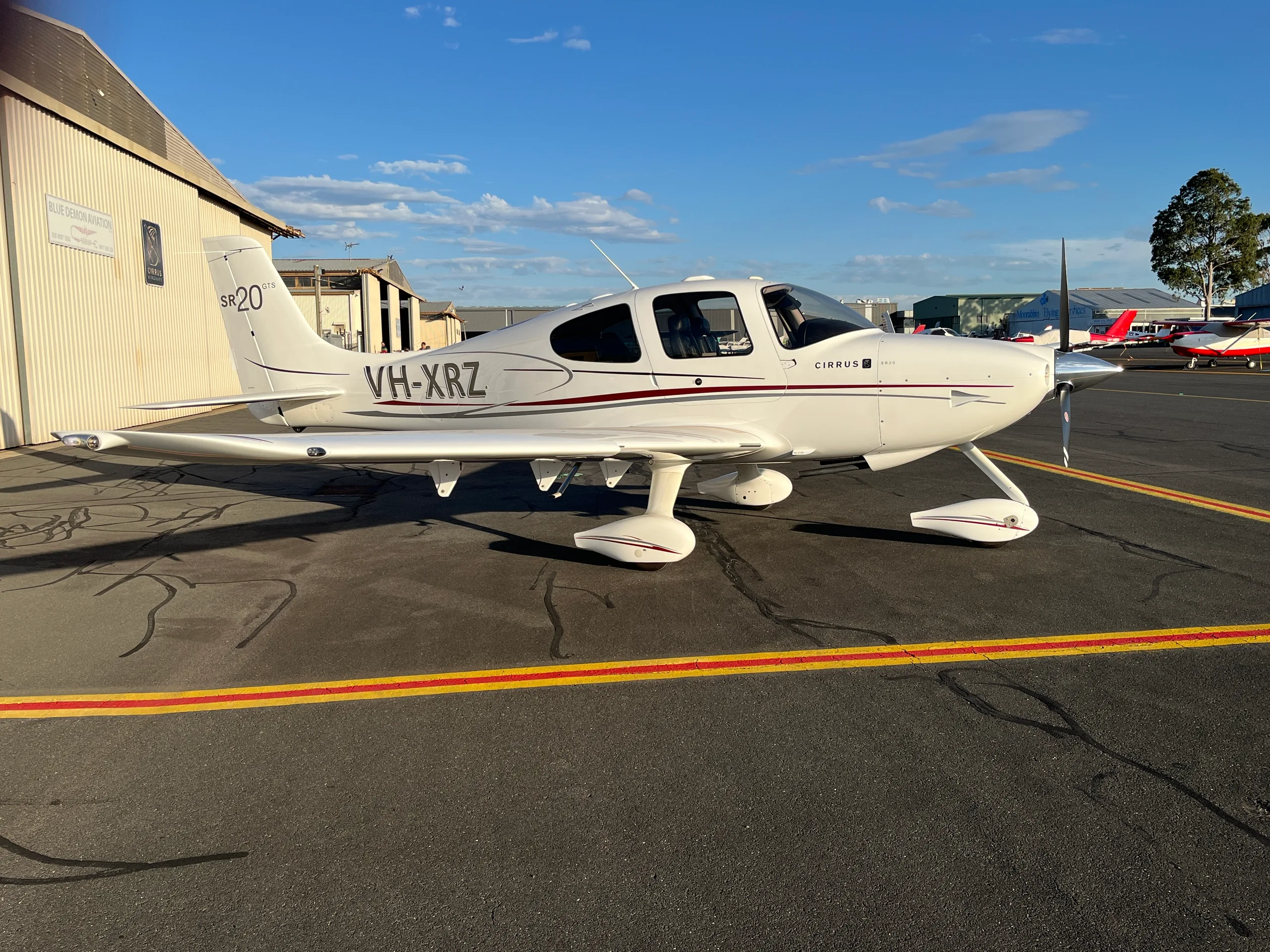 A white Cirrus SR20 GTS aircraft with red and grey stripes, registration VH-XRZ, parked on an airport tarmac next to a hangar.