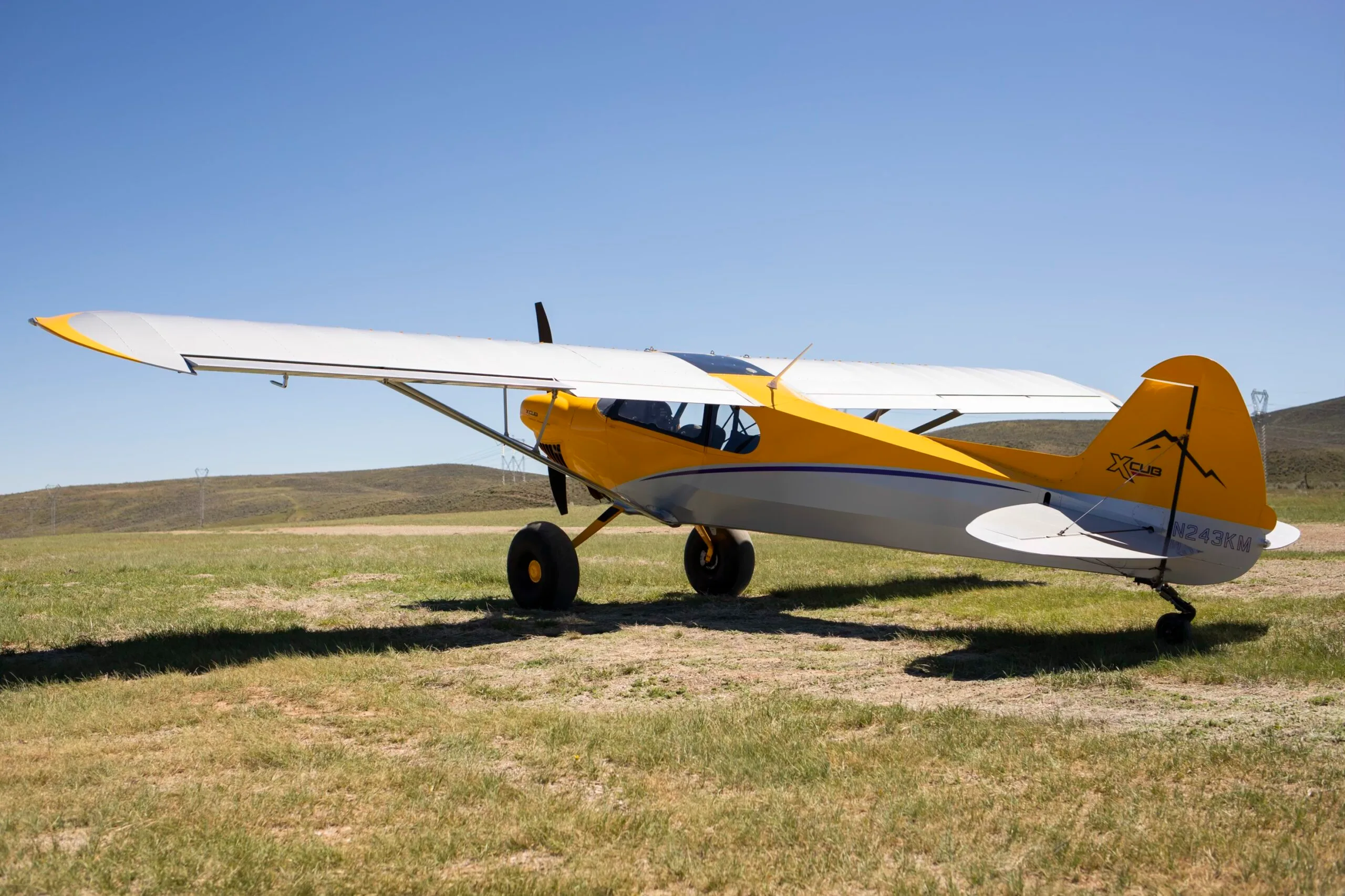 A side view of a yellow and silver CubCrafters XCub aircraft, registration N243KM, parked on a grassy field.