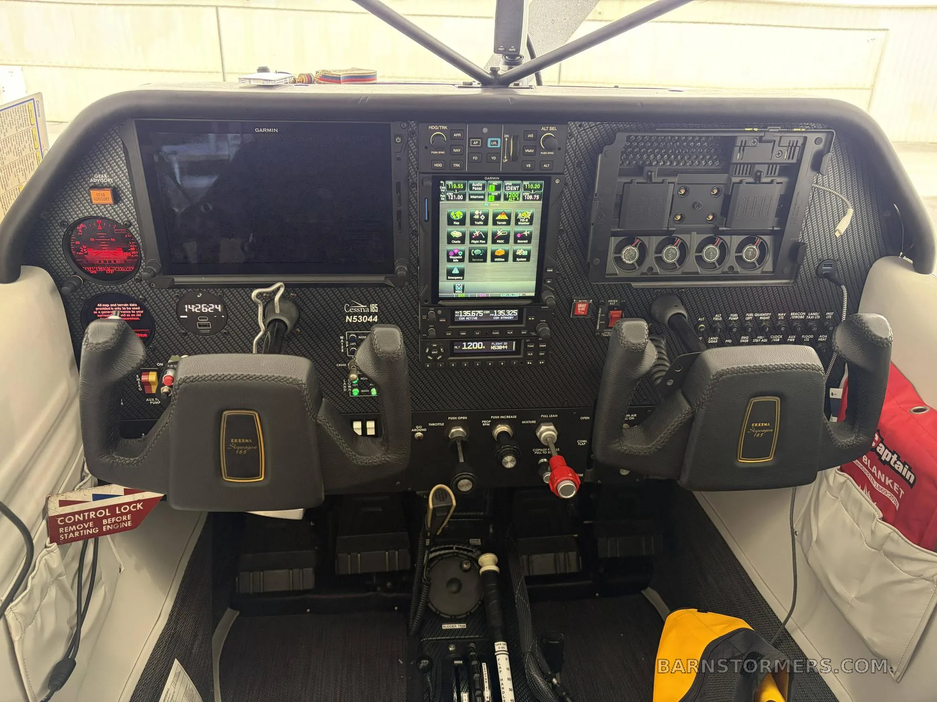 A detailed view of the Cessna 180 Skywagon cockpit, showing the Garmin avionics suite, control yokes, and various switches.