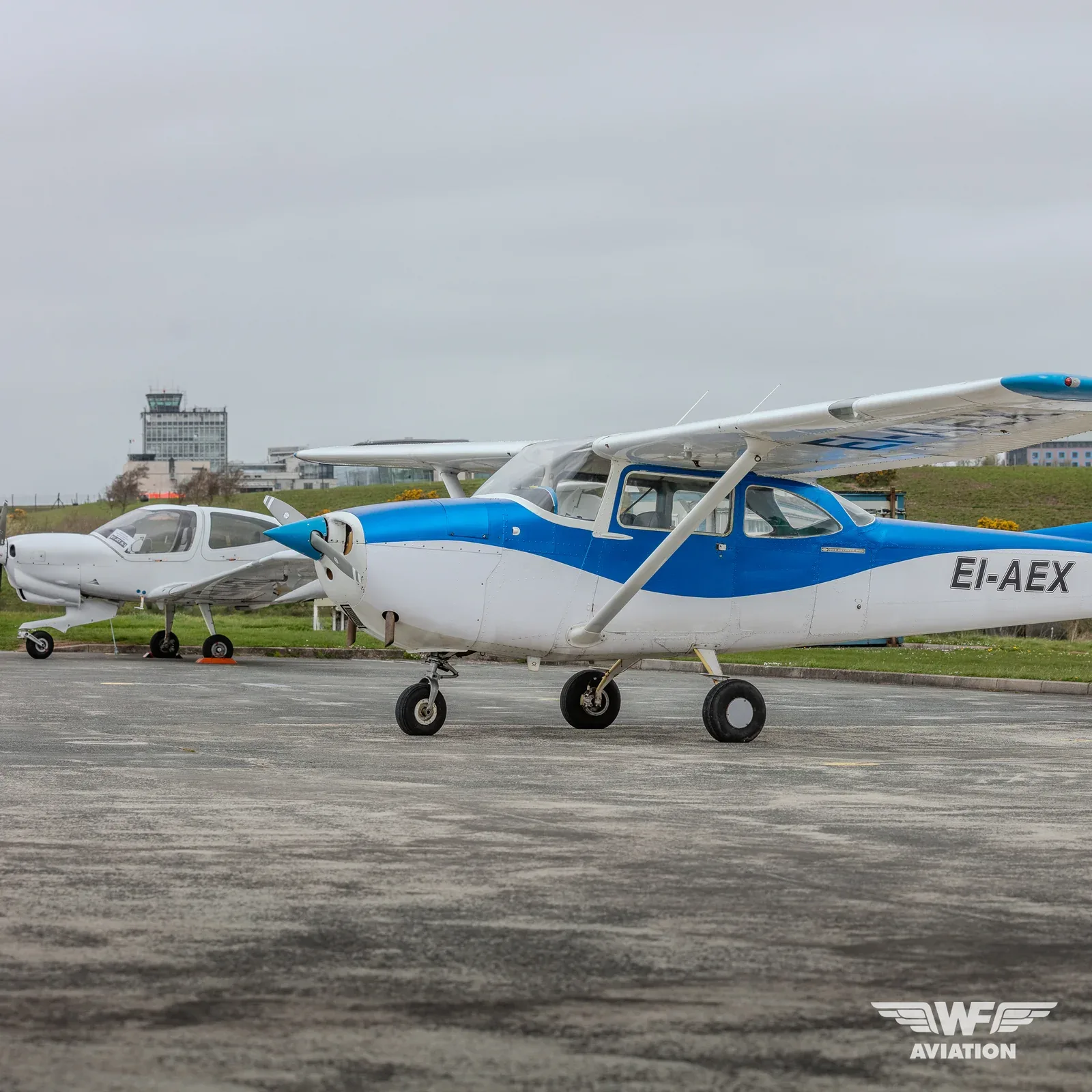 A blue and white Cessna 172-like aircraft is parked on an airfield with another white light aircraft and a control tower in the background.