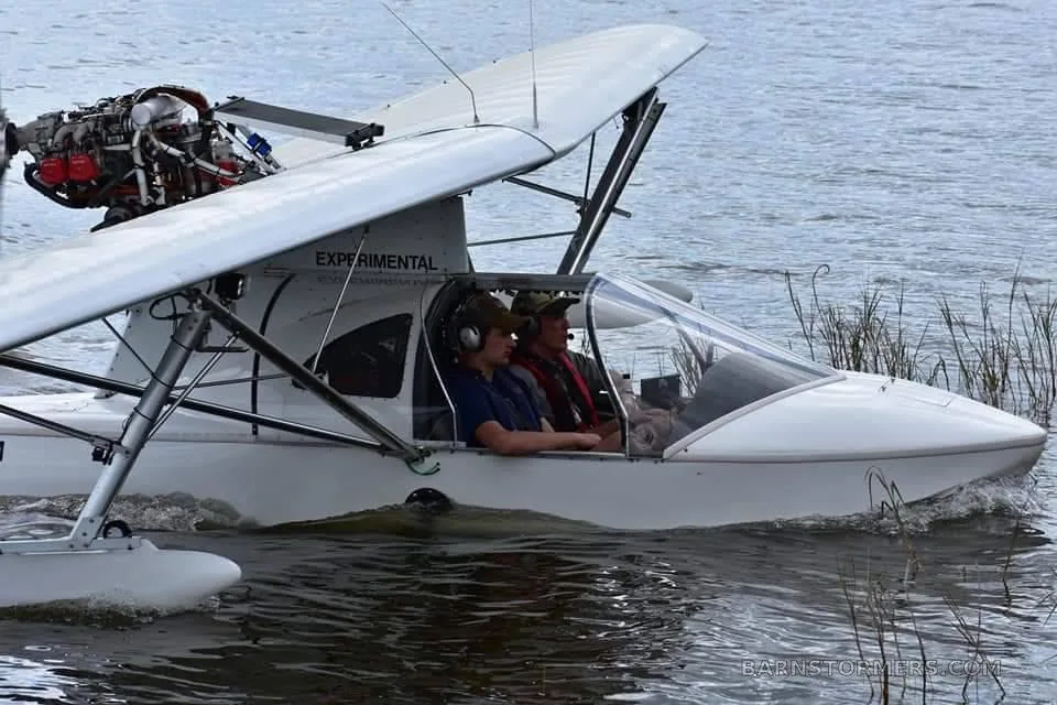 A white experimental seaplane with two people inside is moving on water, viewed from the side.