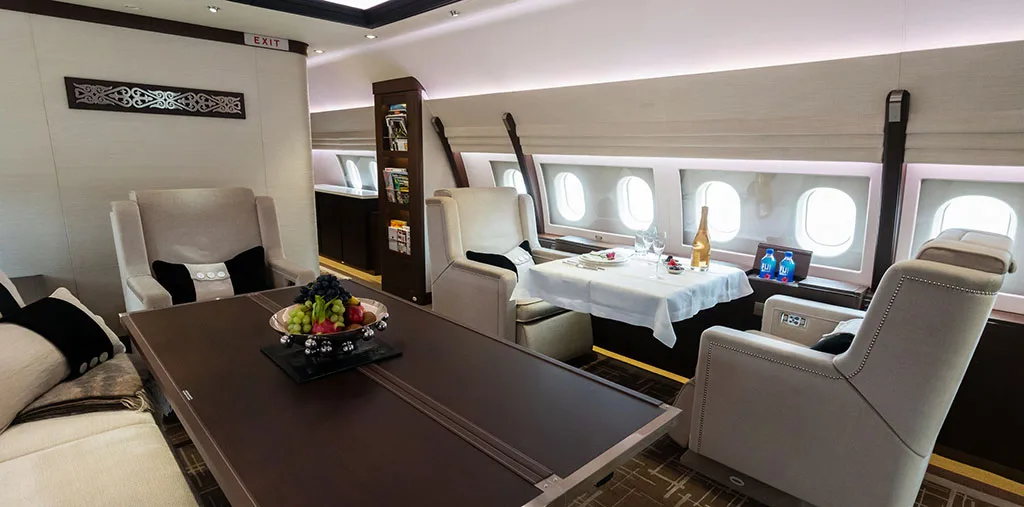 A spacious aircraft cabin featuring a dining table set for two, lounge chairs, and multiple windows.
