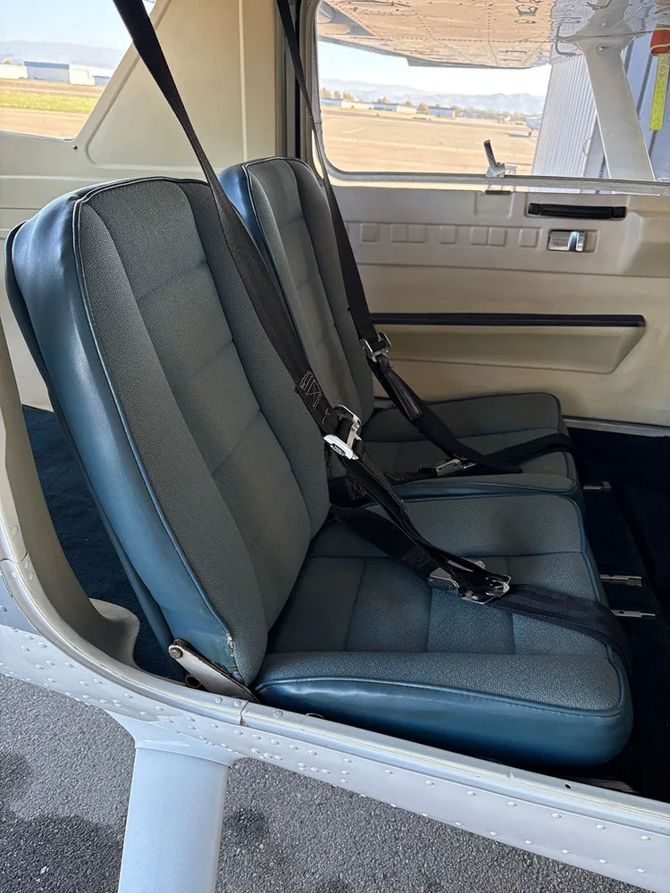 Two blue fabric and vinyl aircraft seats with black seatbelts in the rear cabin, looking out to an airfield.