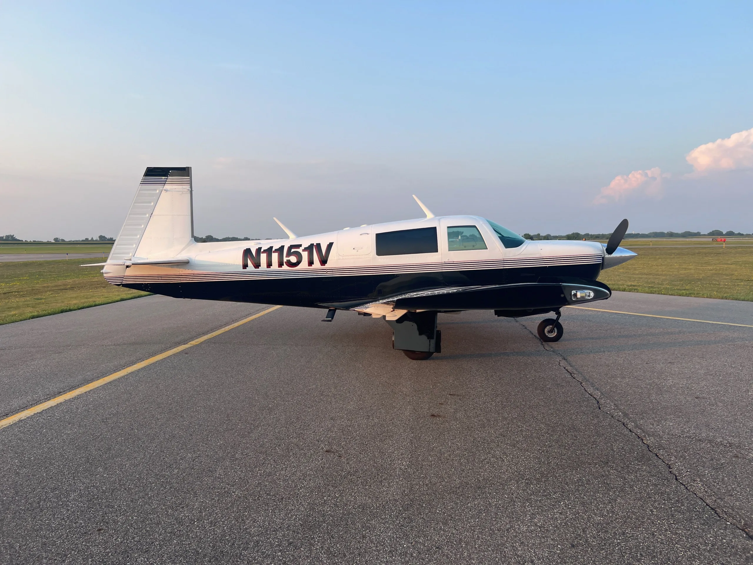 A white and dark blue single-engine aircraft, N1151V, parked on an asphalt runway at sunset.