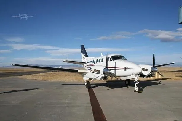 A white twin-engine turboprop aircraft with black and gold stripes is parked on an asphalt runway under a clear blue sky.