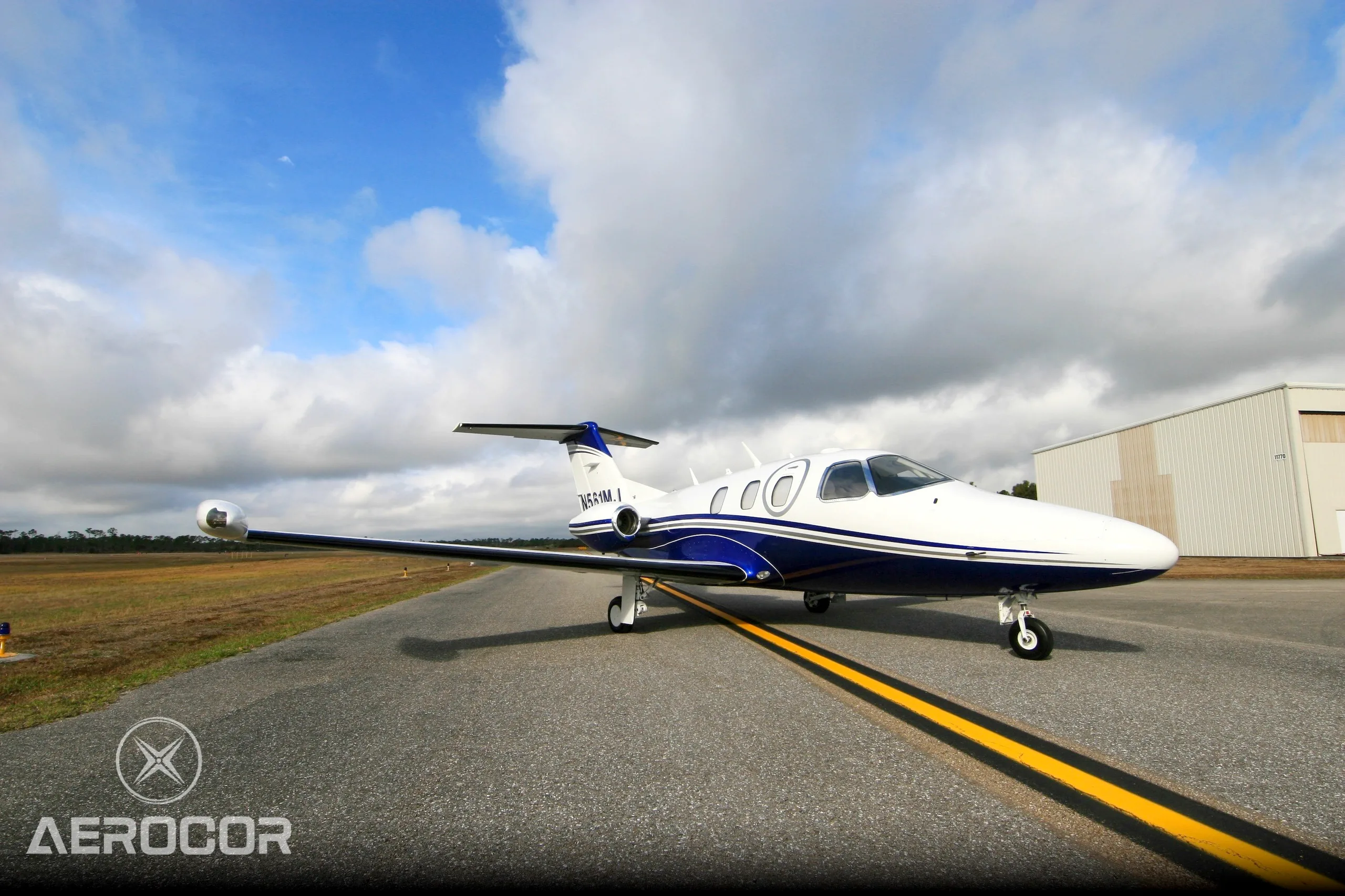 A white and blue Eclipse 500 jet, registration N561MJ, parked on an airfield under a partly cloudy sky.