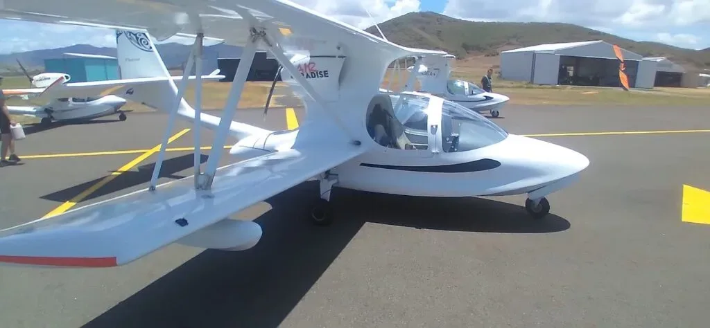 Several white amphibious aircraft are parked on an airfield tarmac with hangars and hills in the background.