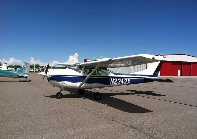 A white and blue Cessna Skylane N2342X parked on an airport tarmac with red hangars in the background.