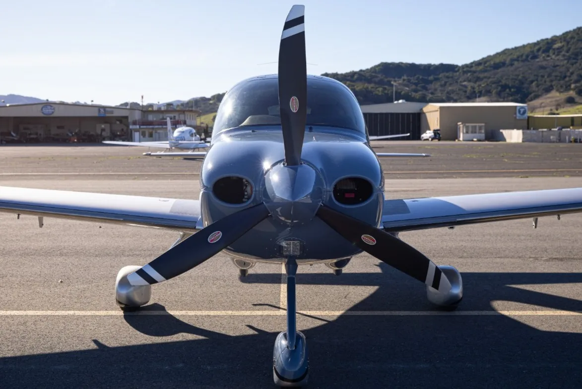 Front view of a light blue single-engine propeller aircraft with a three-blade propeller on an airfield.