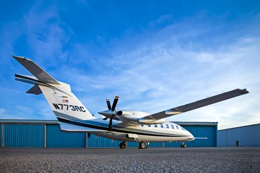 A rear-side view of a white Piaggio P.180 Avanti aircraft with blue and grey stripes, parked on an asphalt tarmac in front of blue hangars.