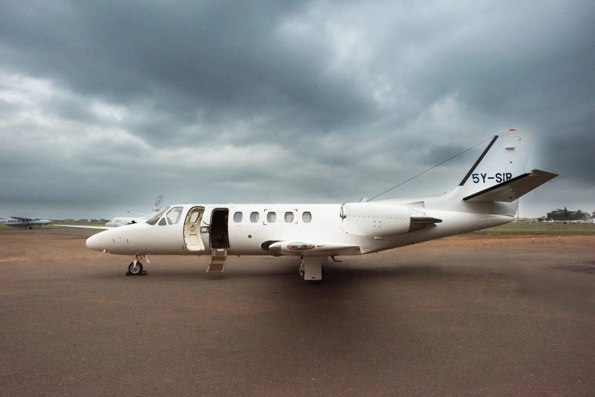 A white Cessna Citation business jet with registration 5Y-SIR parked on a tarmac with its main door open under a cloudy sky.