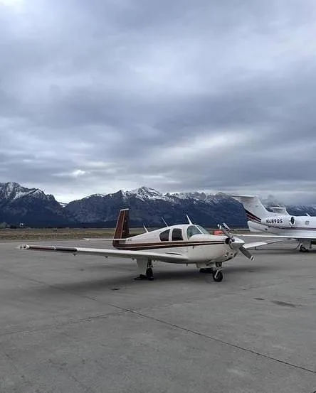 A white and brown single-engine propeller aircraft is parked on an airfield with snow-capped mountains under a cloudy sky.