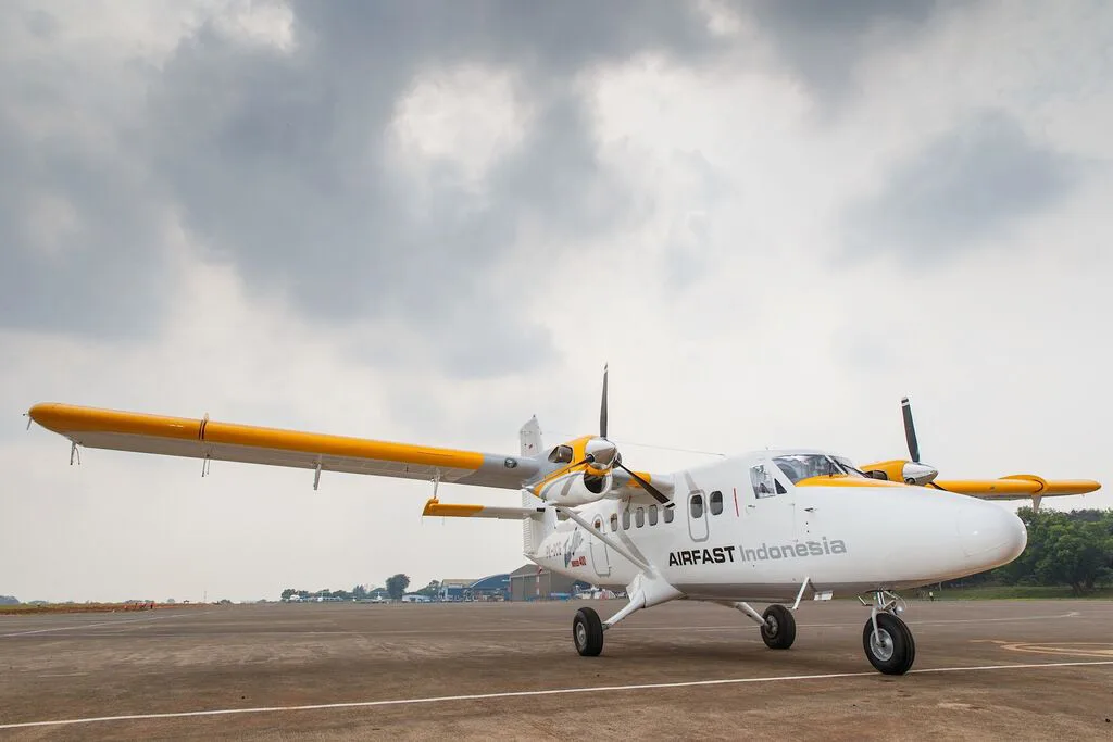 A white and yellow Airfast Indonesia Twin Otter aircraft parked on an airfield under a cloudy sky.