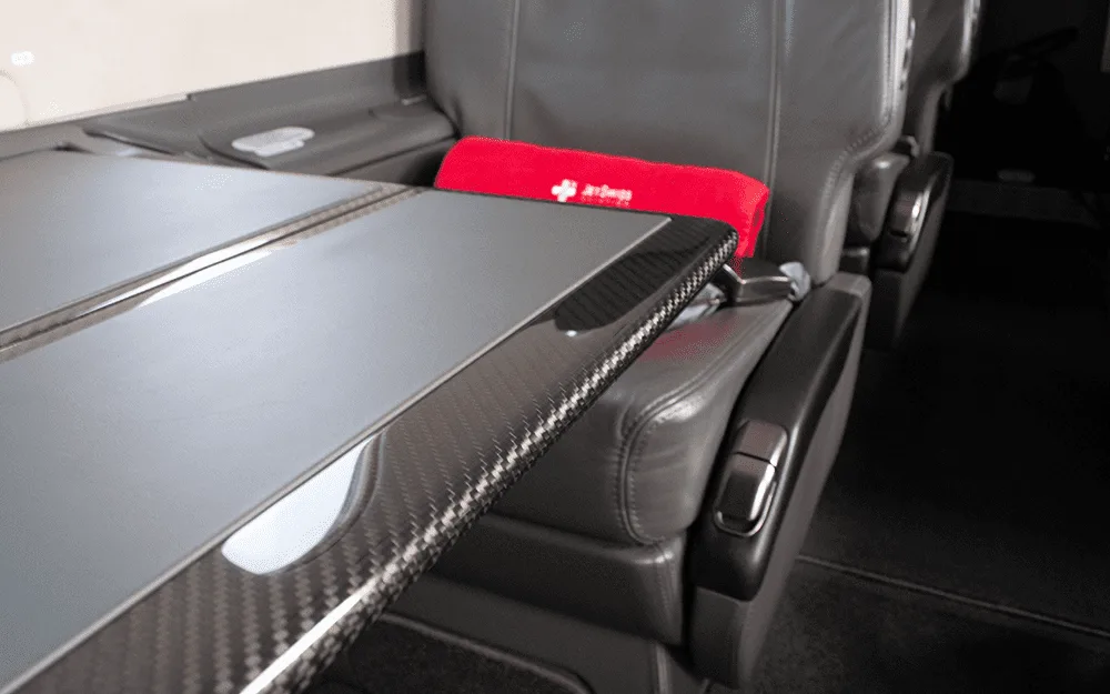 A close-up view of a dark grey leather aircraft seat with a red pillow and a carbon fiber-trimmed fold-out table.