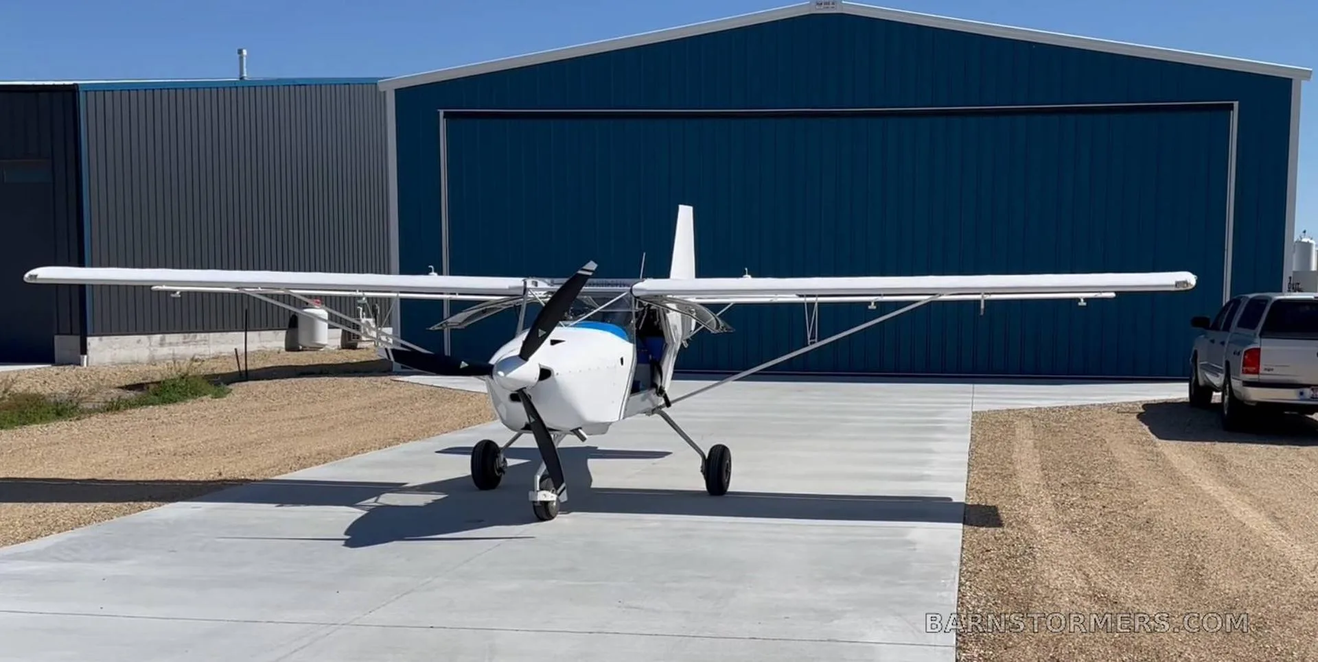 A white Kitfox aircraft with a blue accent, parked on concrete in front of a blue hangar.