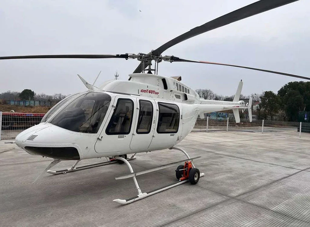 A white Bell 407GXP helicopter with landing skids and ground handling wheels, viewed from the front-left on a concrete pad.