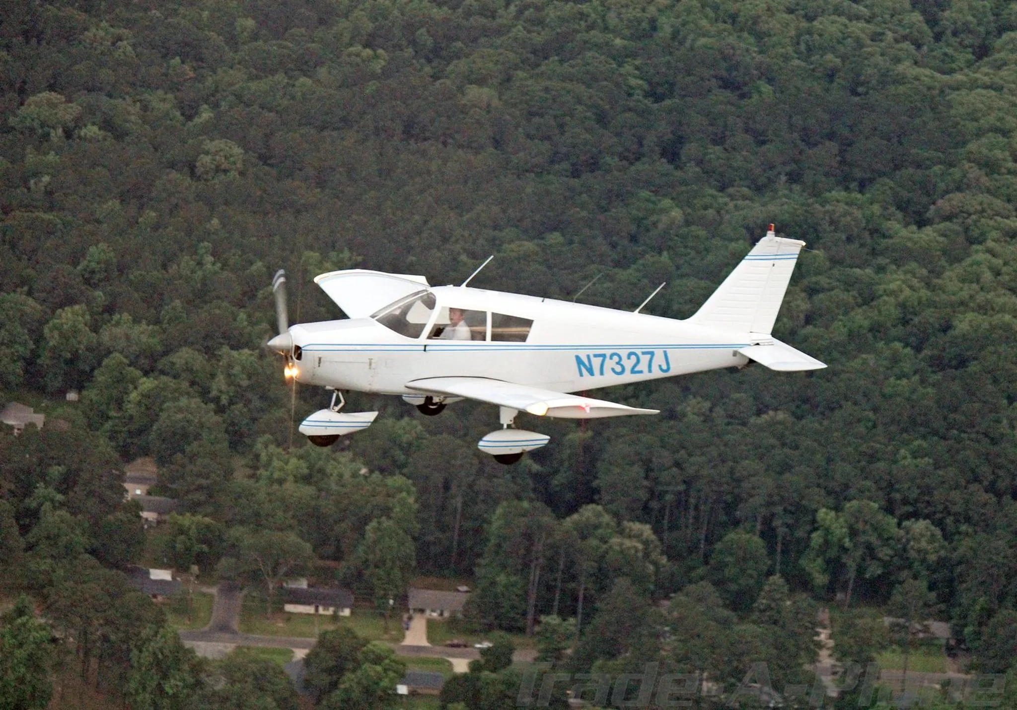 A white Piper Cherokee 140 aircraft, N7327J, with blue stripes, flies over a forested landscape.