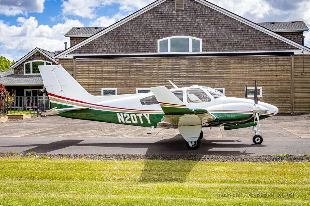 A white and green twin-engine propeller aircraft (N20TY) is parked on a paved surface in front of a large wooden building under a partly cloudy sky.