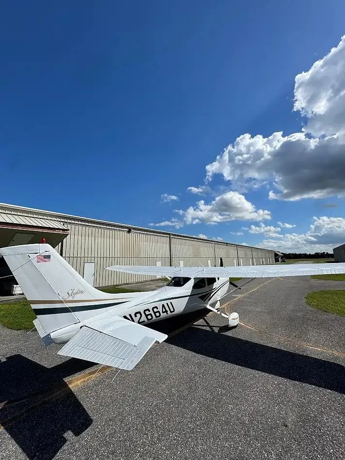 Rear-right view of a white Cessna 182 Skylane with 'N2664U' on the tail, parked on an asphalt ramp next to hangars.
