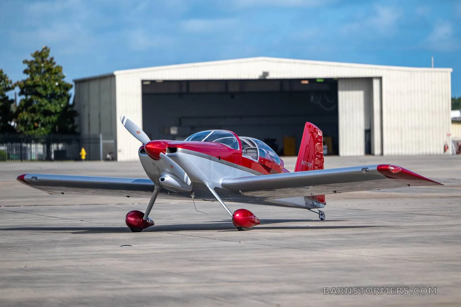A red and silver single-engine experimental aircraft, N711RT, is parked on a tarmac in front of a white hangar.