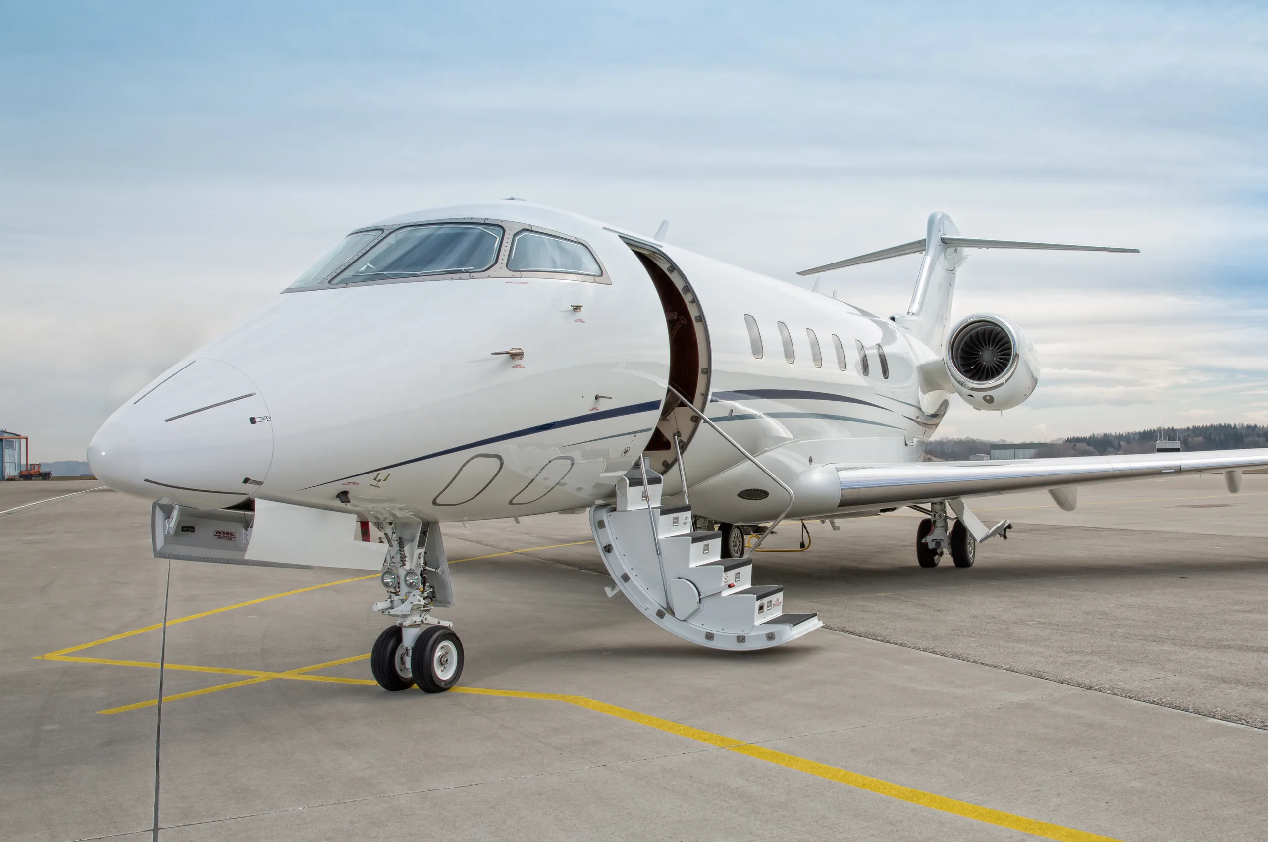 A white business jet with blue accent stripes is parked on an tarmac with its main door open and stairs deployed, under a cloudy sky.