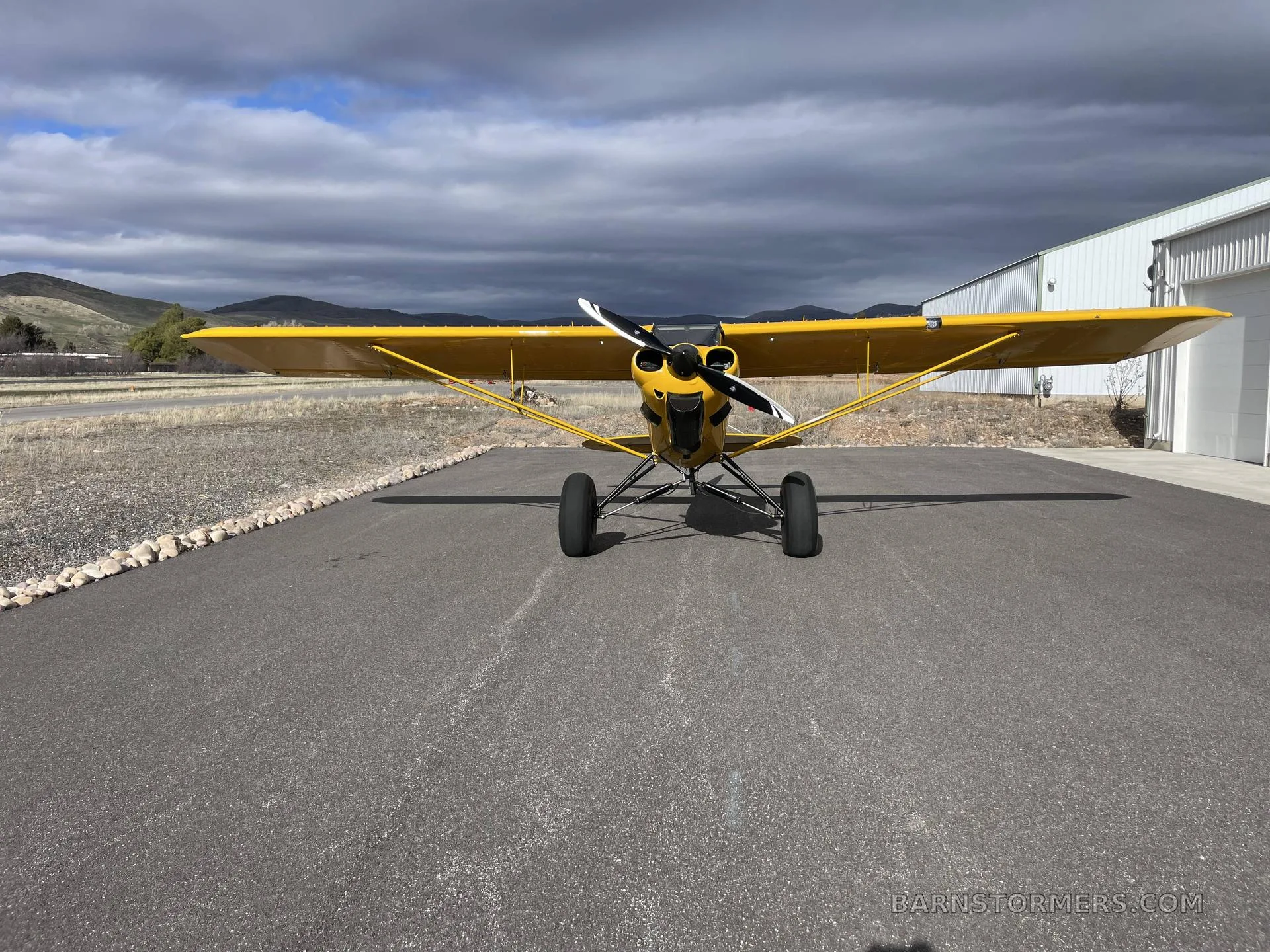 A head-on view of a yellow CubCrafters Carbon Cub EX-2 with a propeller, parked on an asphalt surface in front of a hangar.