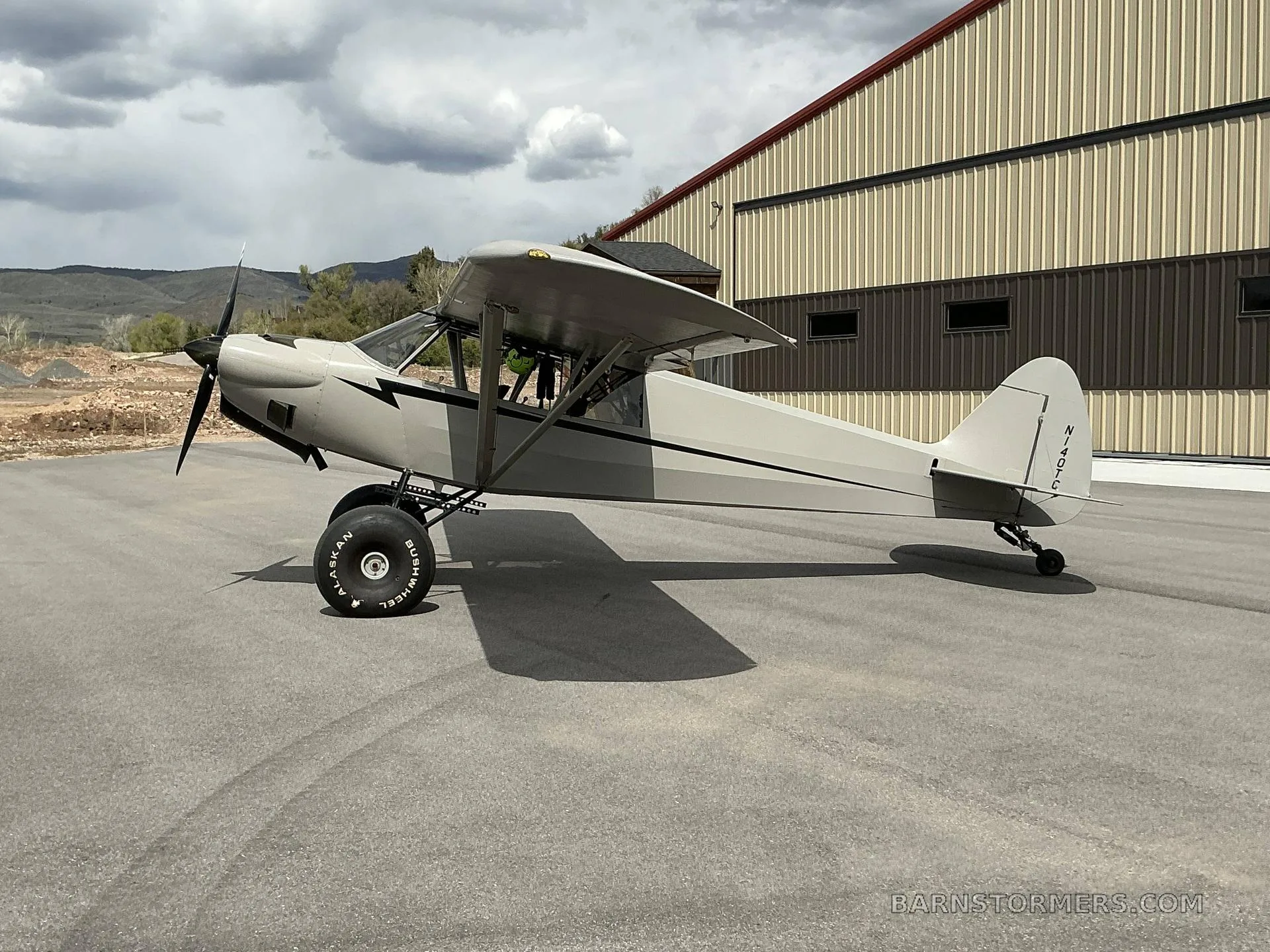 A light grey high-wing taildragger aircraft with black stripes and large tundra tires, parked on asphalt in front of a hangar under a cloudy sky.