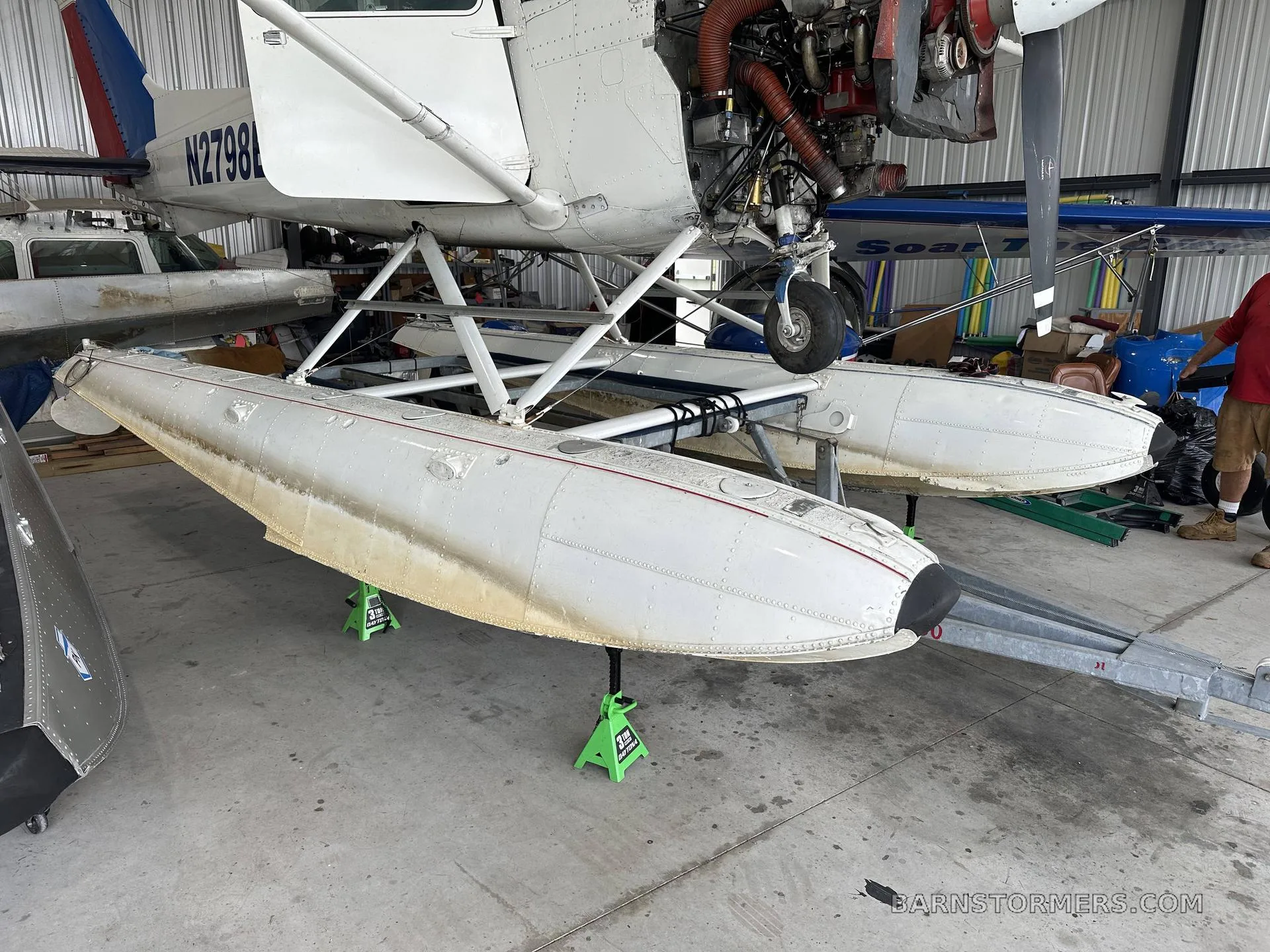 A white floatplane, registration N2798L, is seen on jacks in a hangar, with its engine cowling open.