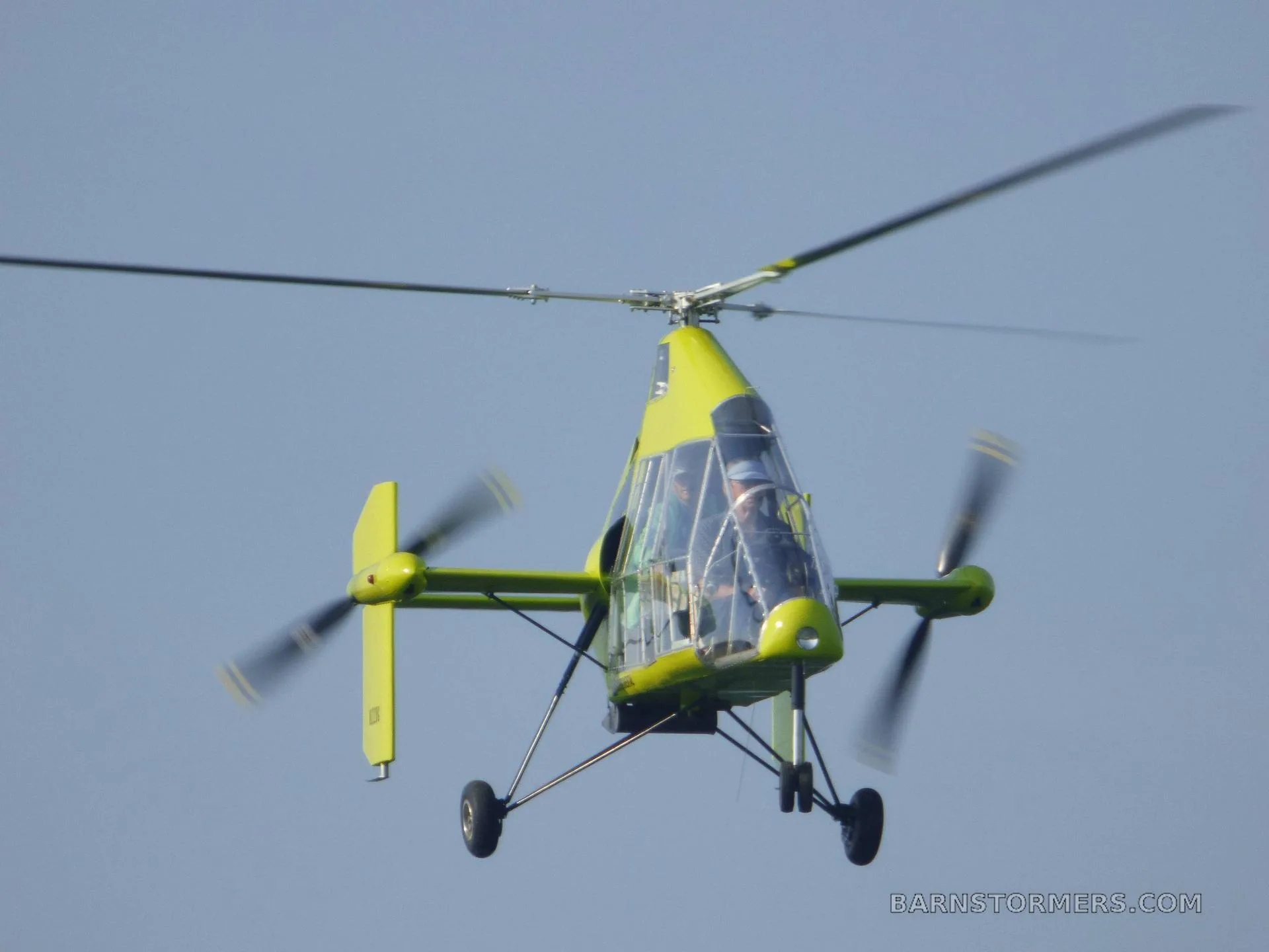 A bright green gyrocopter with a clear canopy and two people inside, flying against a clear blue sky.