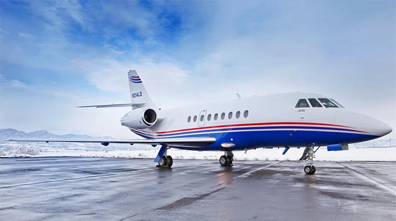 A Dassault Falcon private jet with a white, blue, and red livery is parked on a wet tarmac with snowy mountains in the background.
