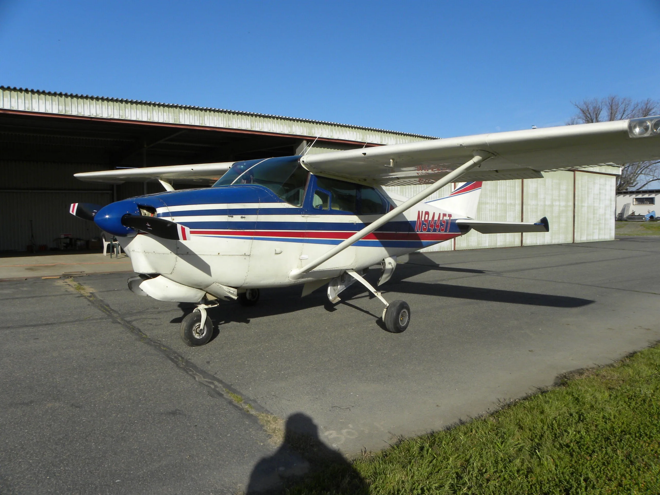 A white Cessna 182 with blue and red stripes, registration N9445T, parked on a tarmac.