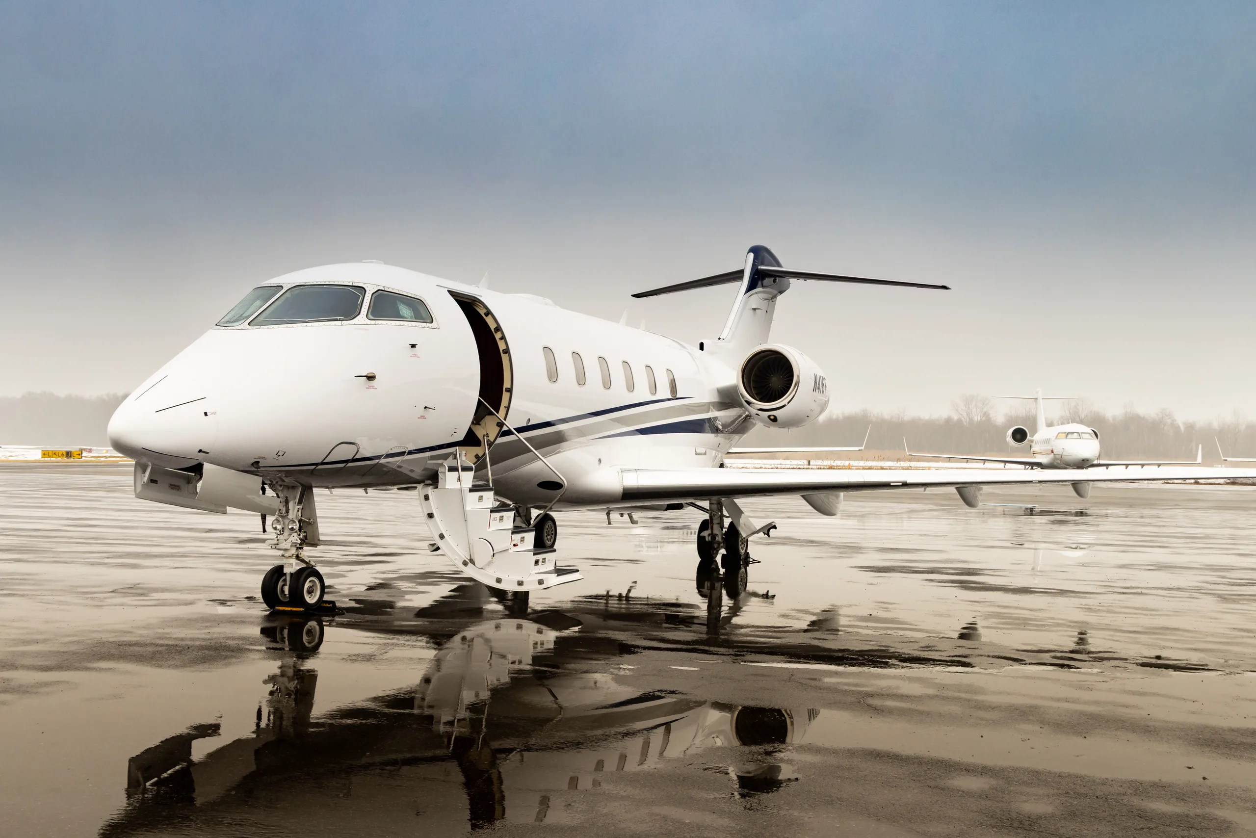A white Bombardier Challenger 300 business jet with blue and grey stripes on a wet tarmac, with its main door open.