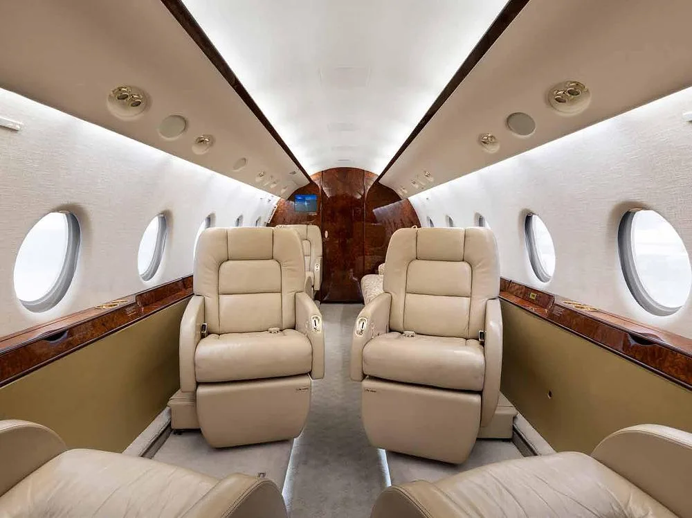 The luxurious interior cabin of a private jet with beige leather seats, wood paneling, and large oval windows, viewed from the rear.