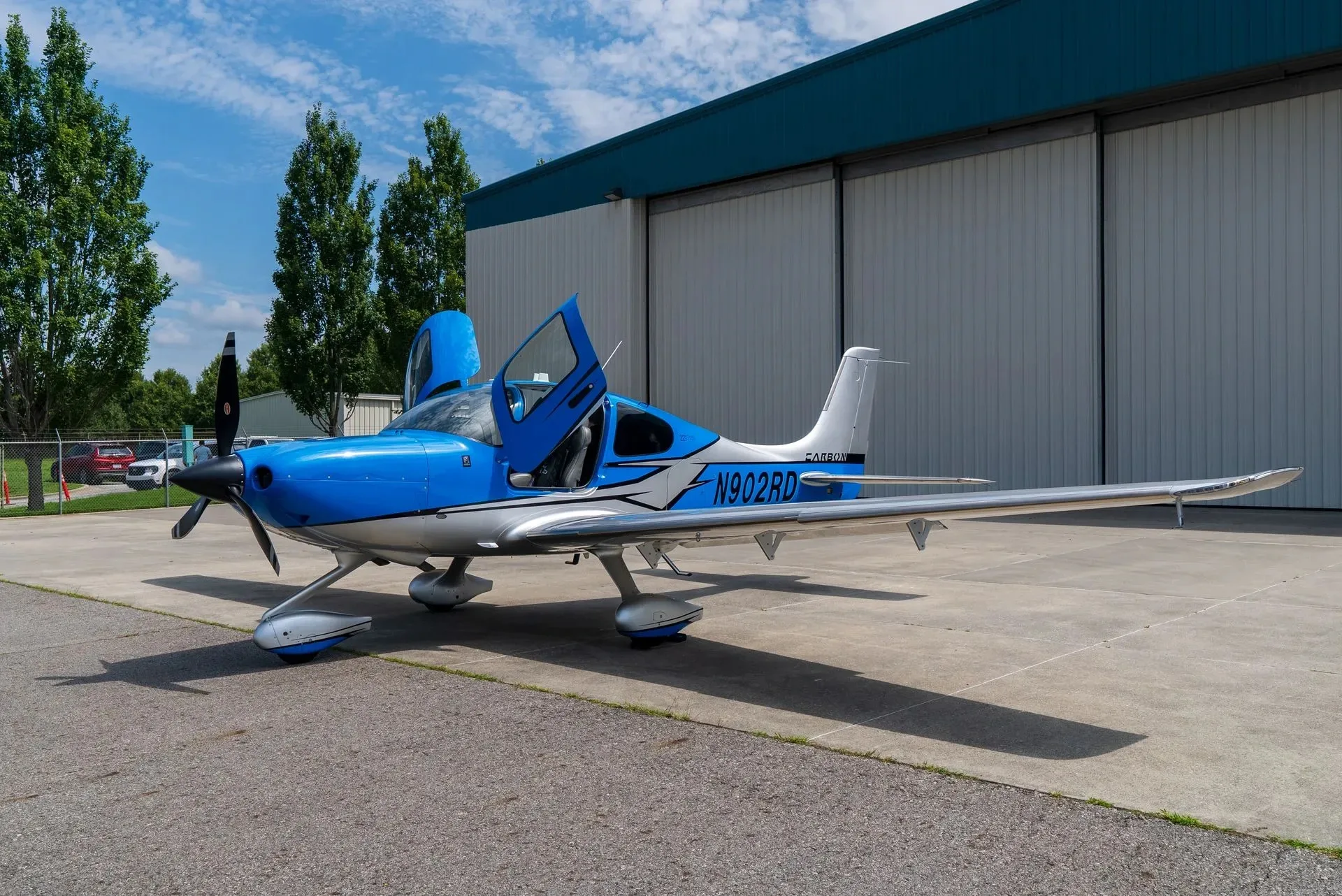 A blue and silver Cirrus SR22 aircraft with its doors open, parked on an airfield in front of a hangar.