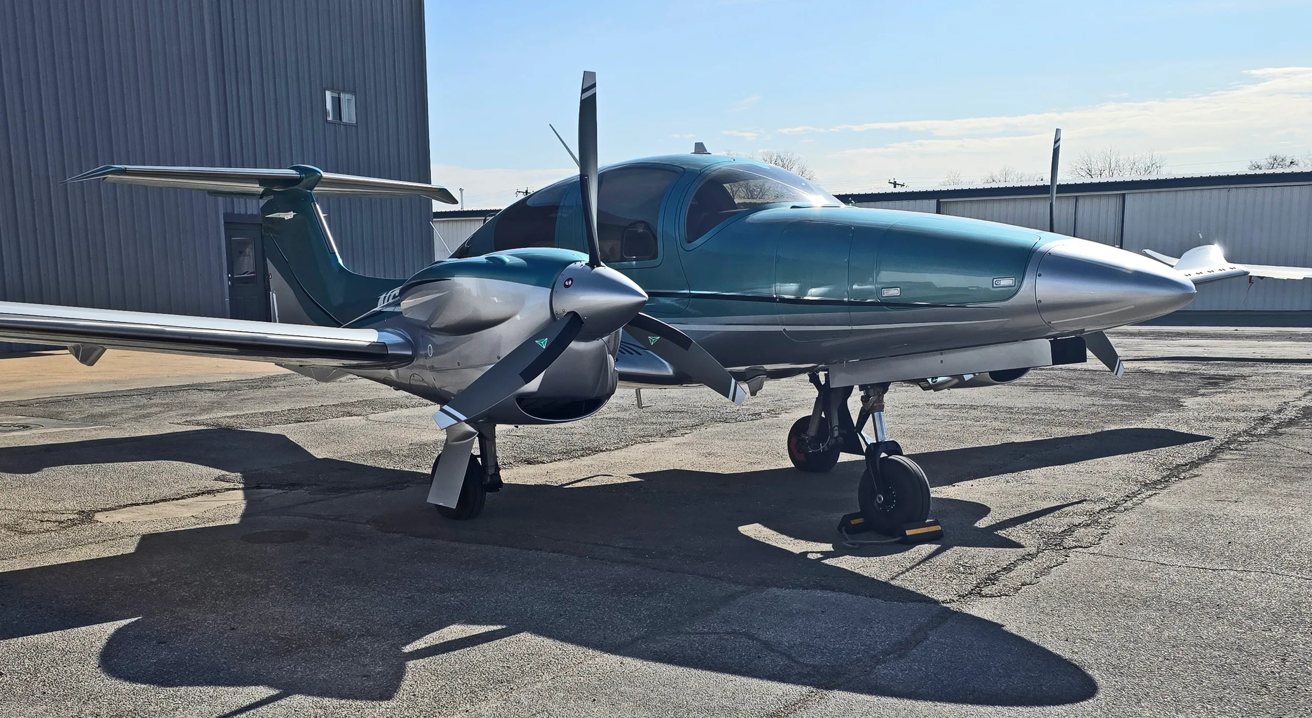 A side view of a teal and silver Diamond DA62 twin-engine aircraft parked on an asphalt ramp under a blue sky.