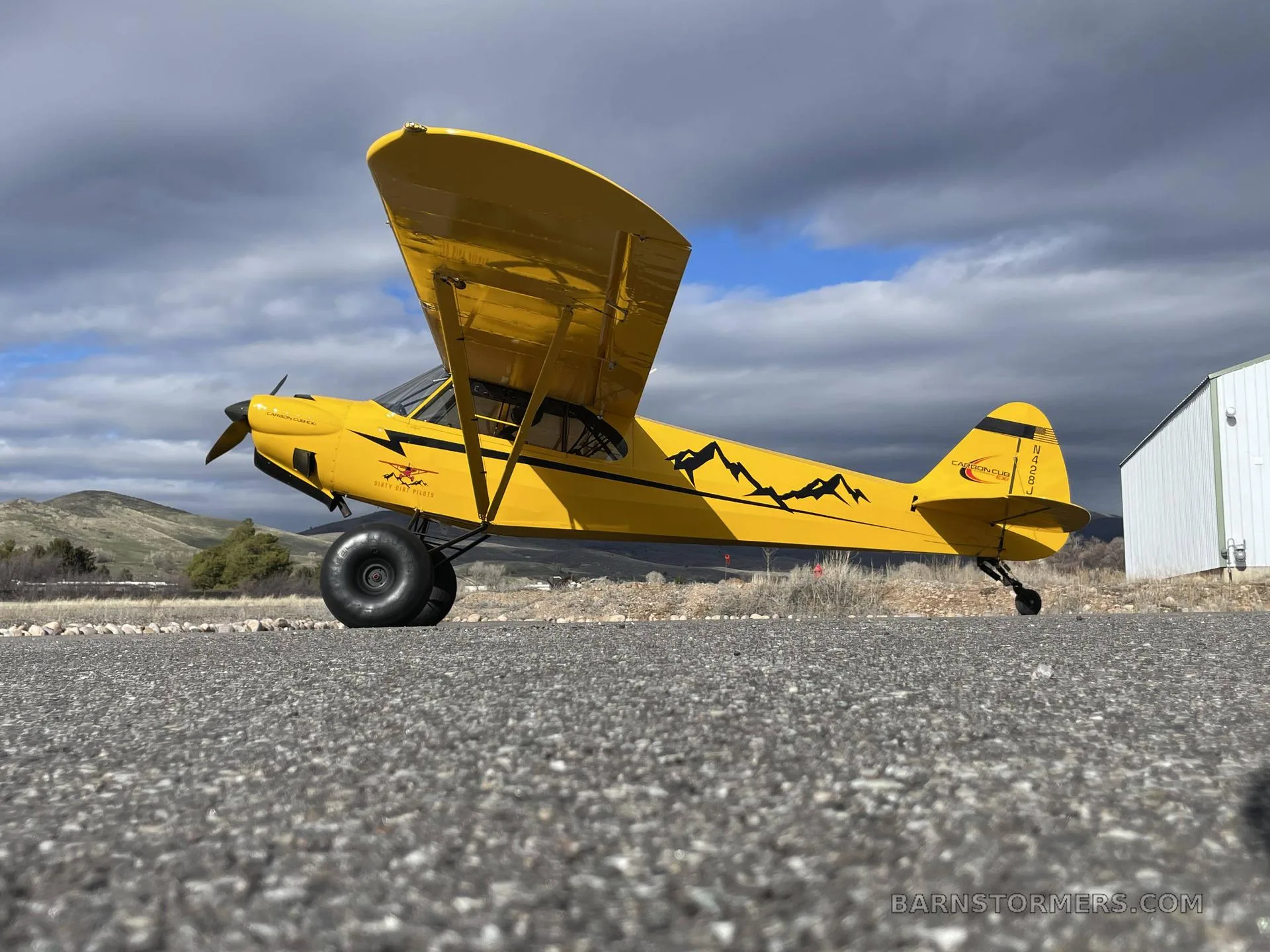 A yellow CubCrafters Carbon Cub EX-2 with large tundra tires and mountain decals, parked on an asphalt runway under a cloudy sky.