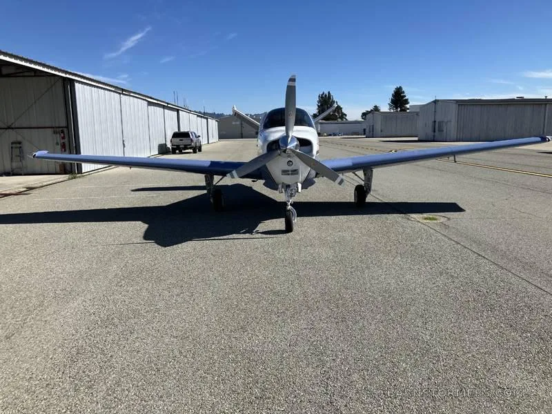 A front view of a light blue and white single-engine aircraft with a propeller, parked on an asphalt tarmac.