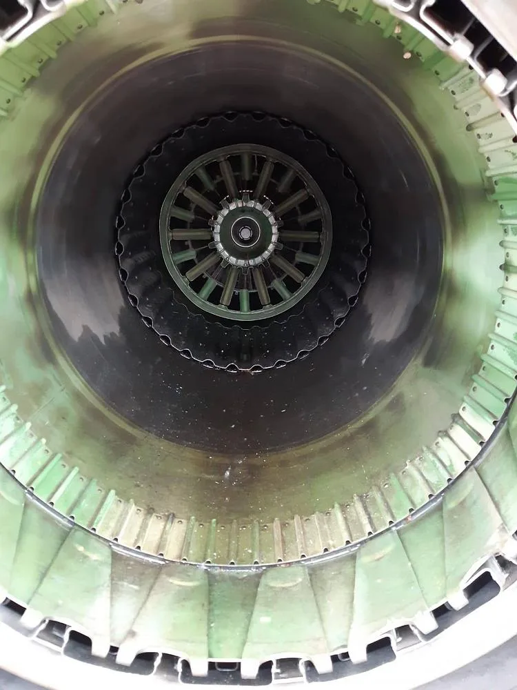 A close-up view looking into the green-tinted exhaust nozzle of a jet engine, showing the turbine blades.