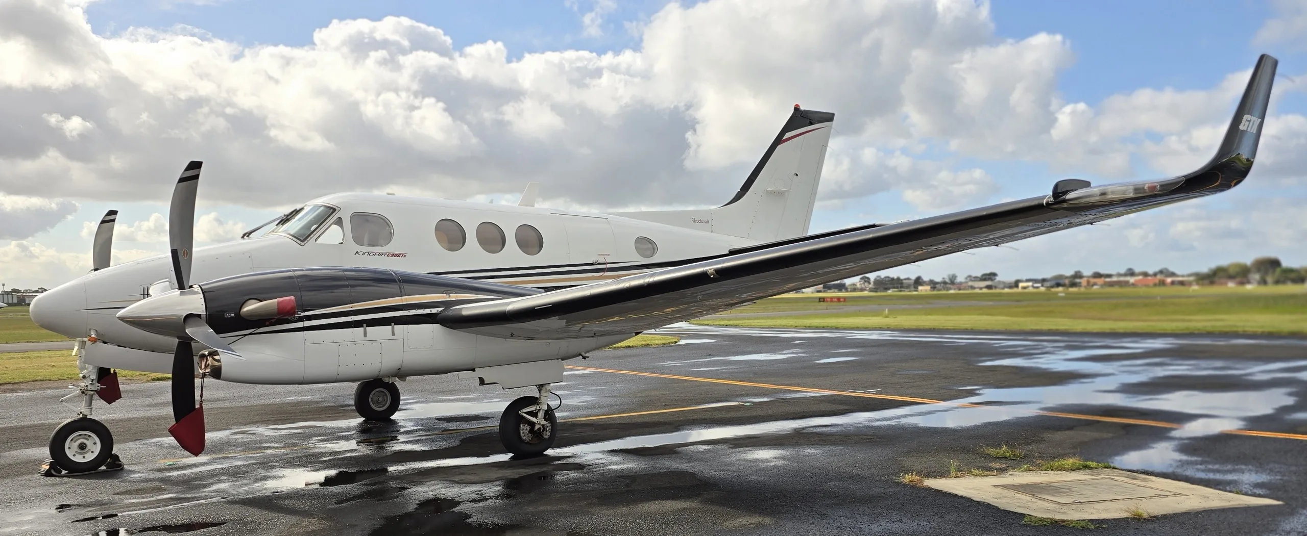 A white and black Beechcraft King Air 200 with gold stripes parked on a wet tarmac under a cloudy sky.
