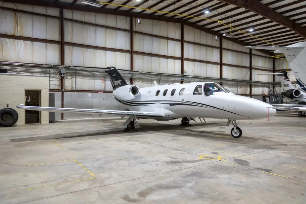 A white Cessna Citation M2 business jet with black and grey stripes is parked inside a hangar.