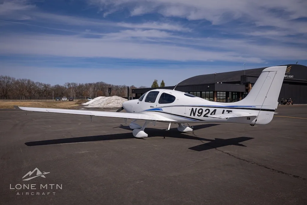 A white single-engine aircraft with blue and black stripes, tail number N924JT, parked on an airport tarmac with a hangar in the background.