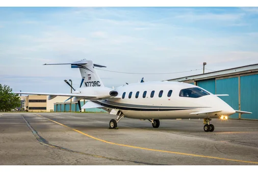 A front-side view of a white Piaggio P.180 Avanti aircraft with blue and grey stripes, parked on an asphalt tarmac with buildings in the background.