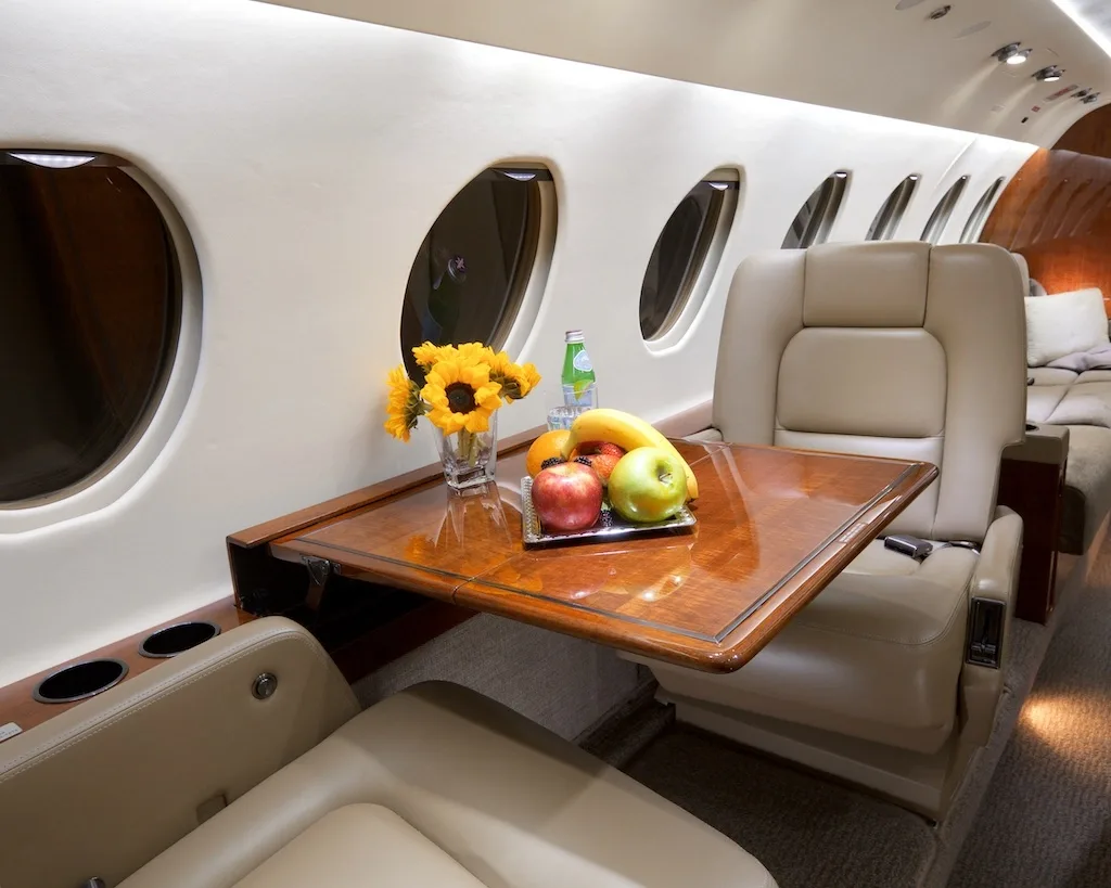 A close-up of a polished wooden table in a private jet cabin, laden with fresh fruit, a bottle of water, and a vase of sunflowers.