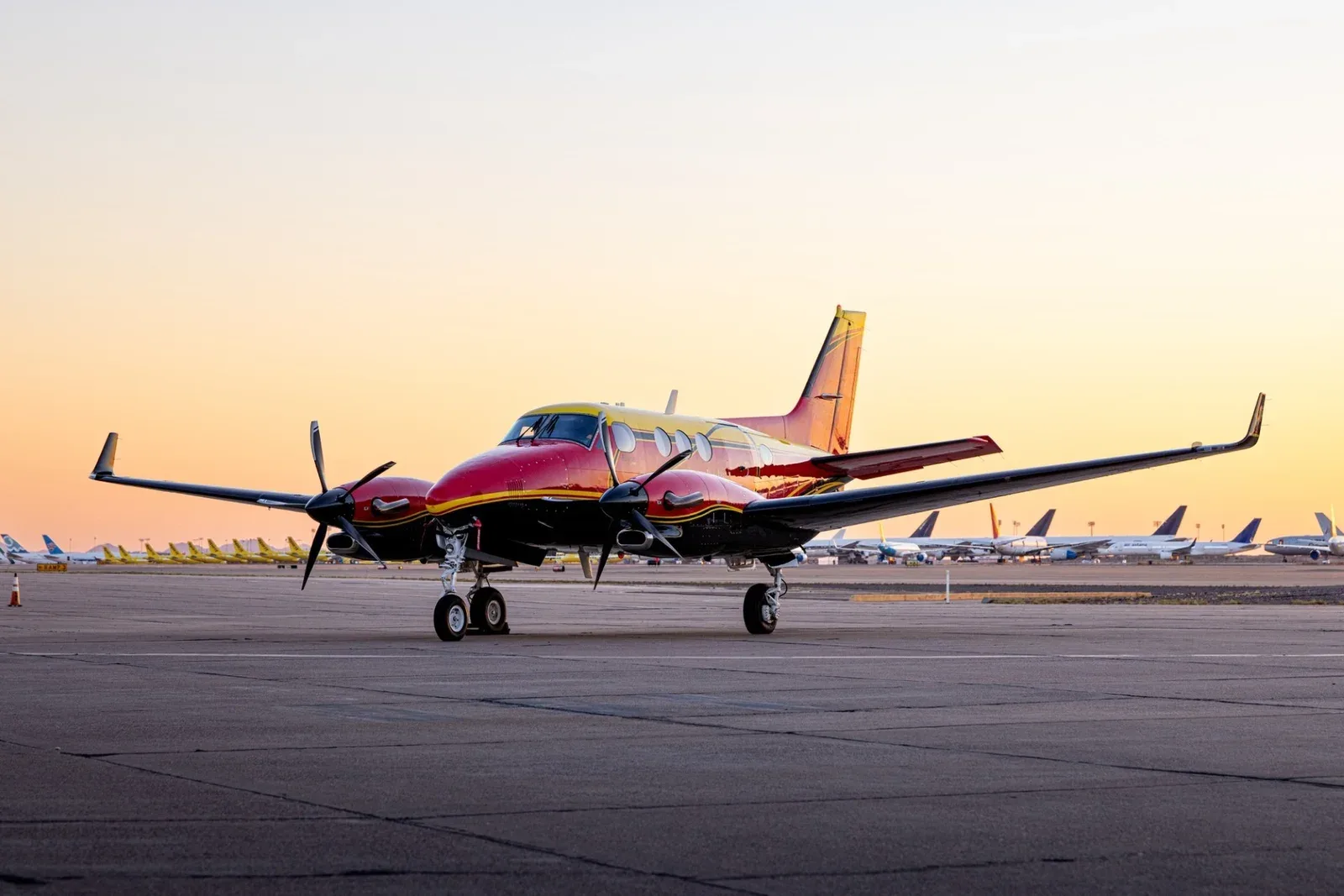 A red, yellow, and black King Air C90B turboprop aircraft parked on an airfield at sunset.