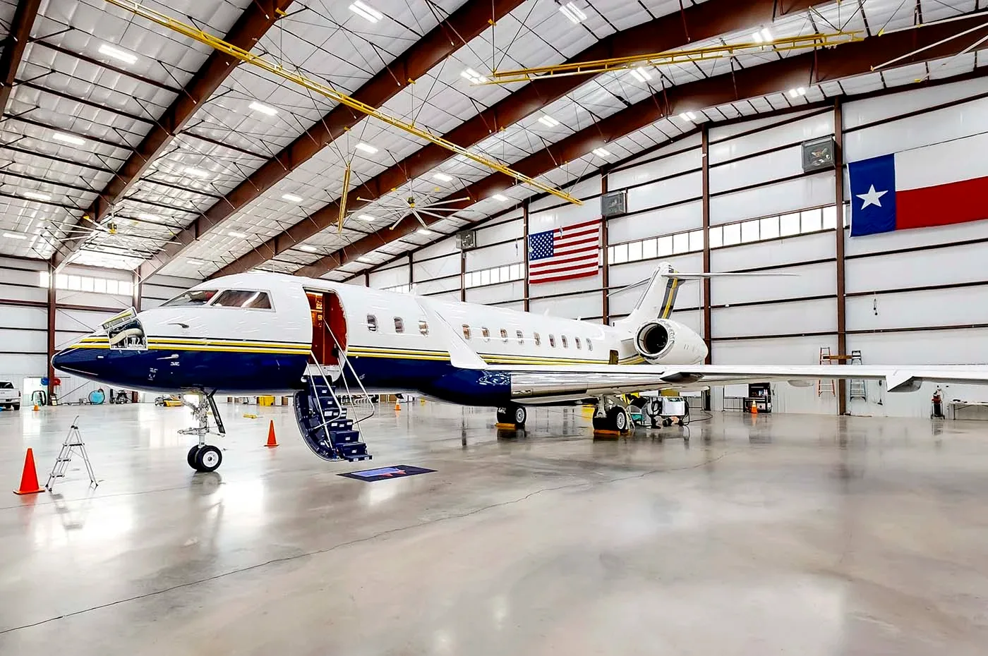 A large white business jet with a blue and gold stripe parked inside a hangar with US and Texas flags.