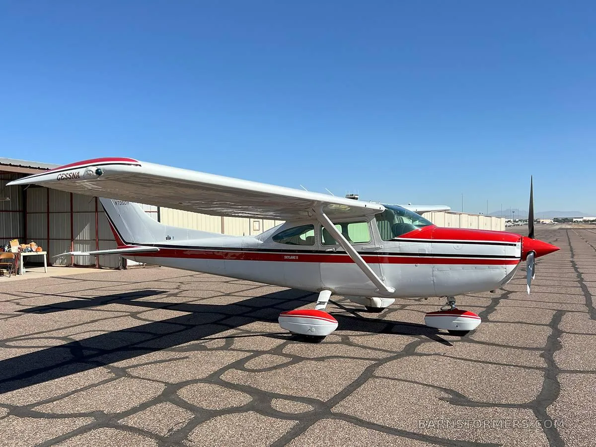A white and red Cessna Skylane II aircraft is parked on an asphalt tarmac with a hangar in the background.