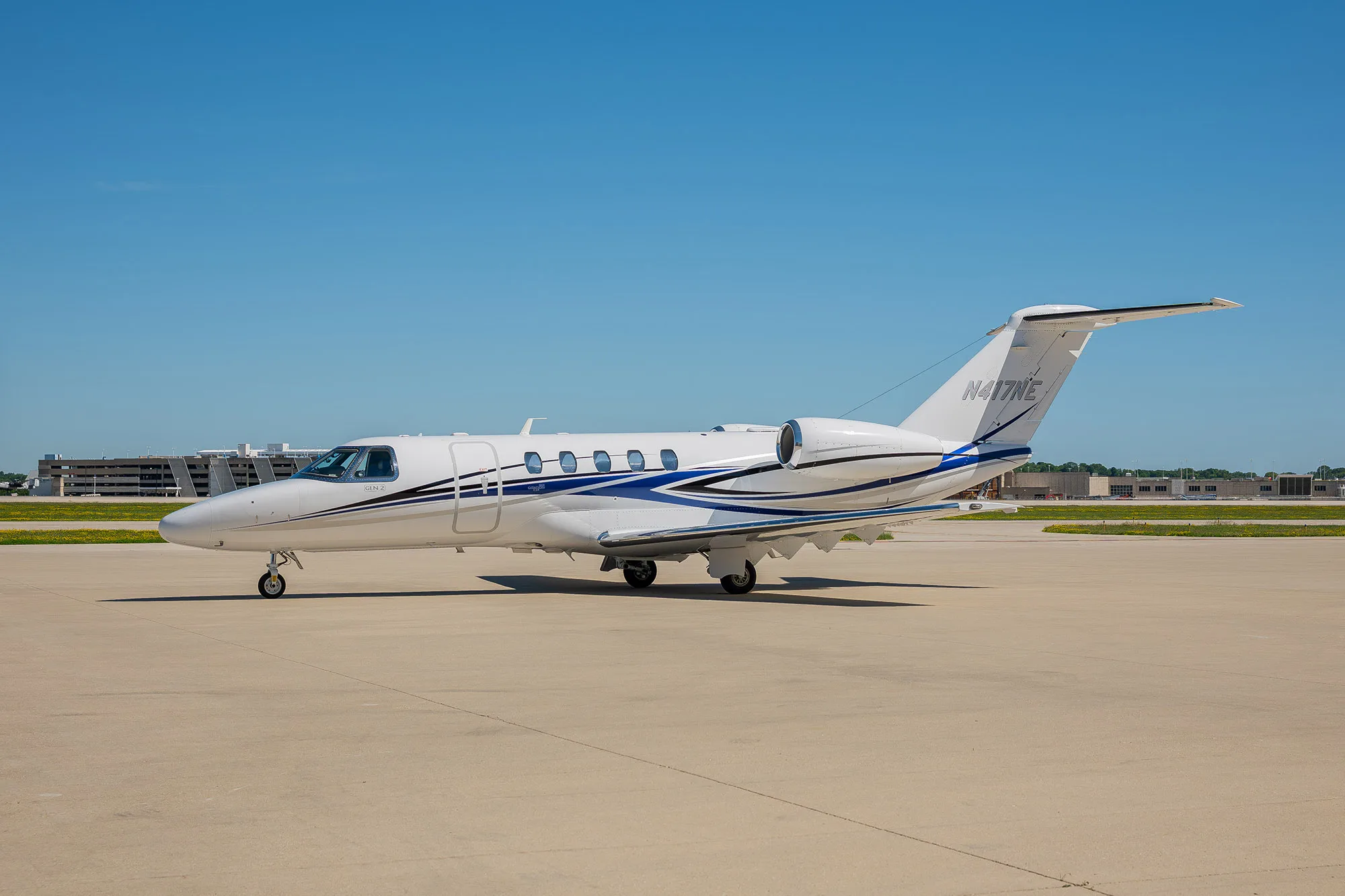 A white business jet with blue and grey stripes parked on an airfield under a clear blue sky.