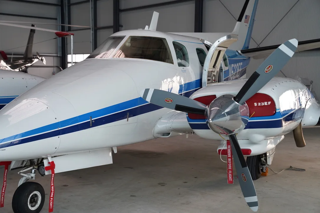 A white twin-engine propeller aircraft with blue stripes, parked in a hangar with its passenger door open.