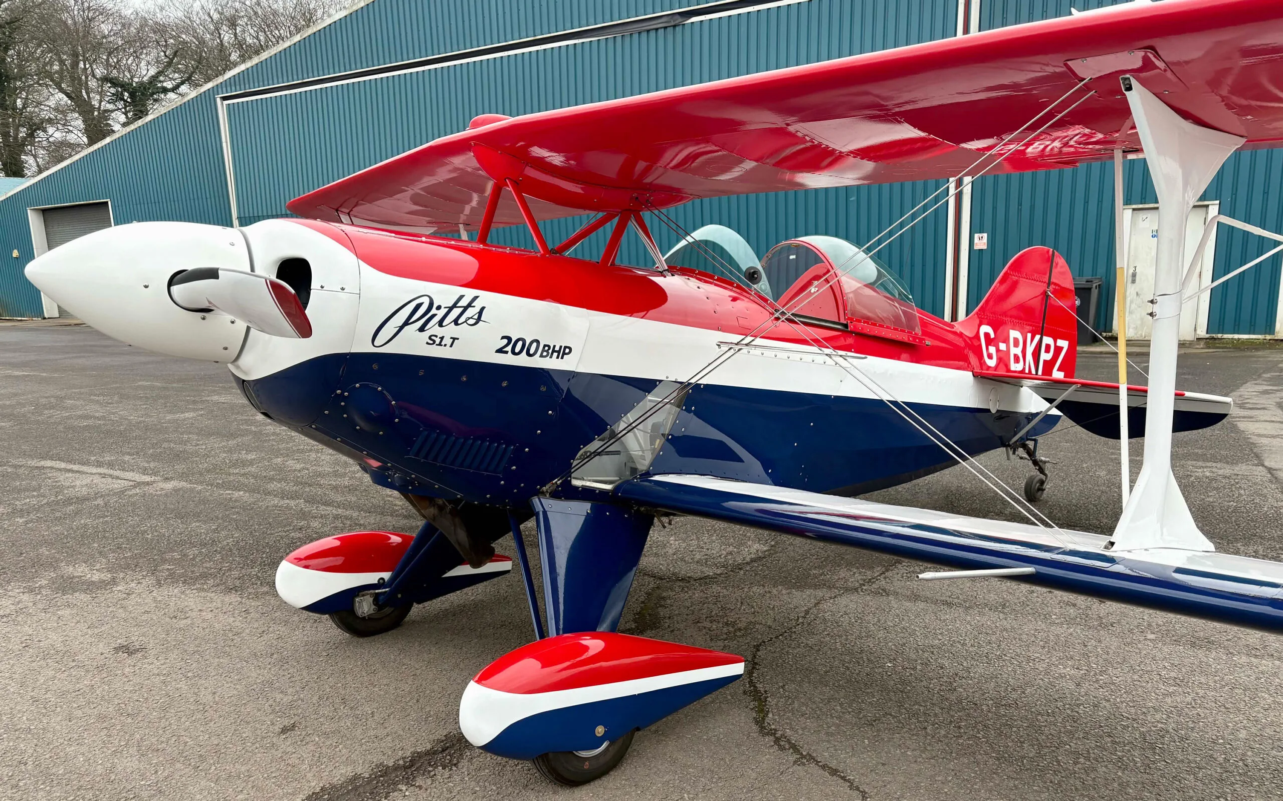 A red, white, and blue Pitts S1T biplane (G-BKPZ) parked on an airfield tarmac next to a hangar.